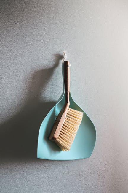 A light blue dustpan hanging on a plain, white wall, holding a wooden-handled broom with beige bristles. The dustpan and broom are positioned vertically, casting a shadow on the wall behind them. The 