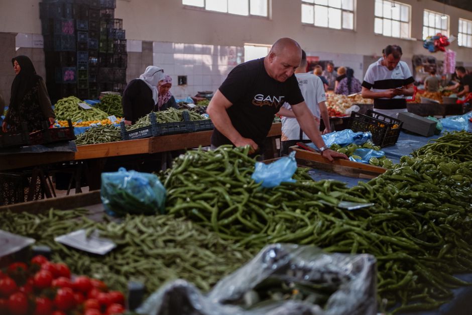 A man wearing a dark brown beanie and navy jacket is shopping at a fruit stall in a market. The stall displays fresh, brightly colored vegetables including red tomatoes, green beans, and zucchini, arr