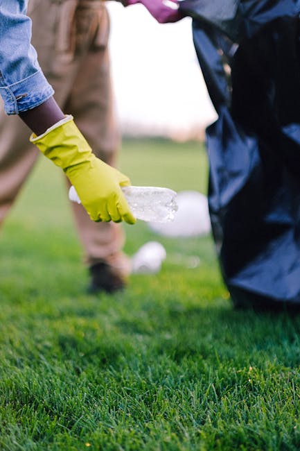 Close-up of a person wearing a neon yellow glove using a clear spray bottle to disinfect or clean a grassy surface in Brockwell Park, SW2, with another individual nearby wearing a dark raincoat. The b